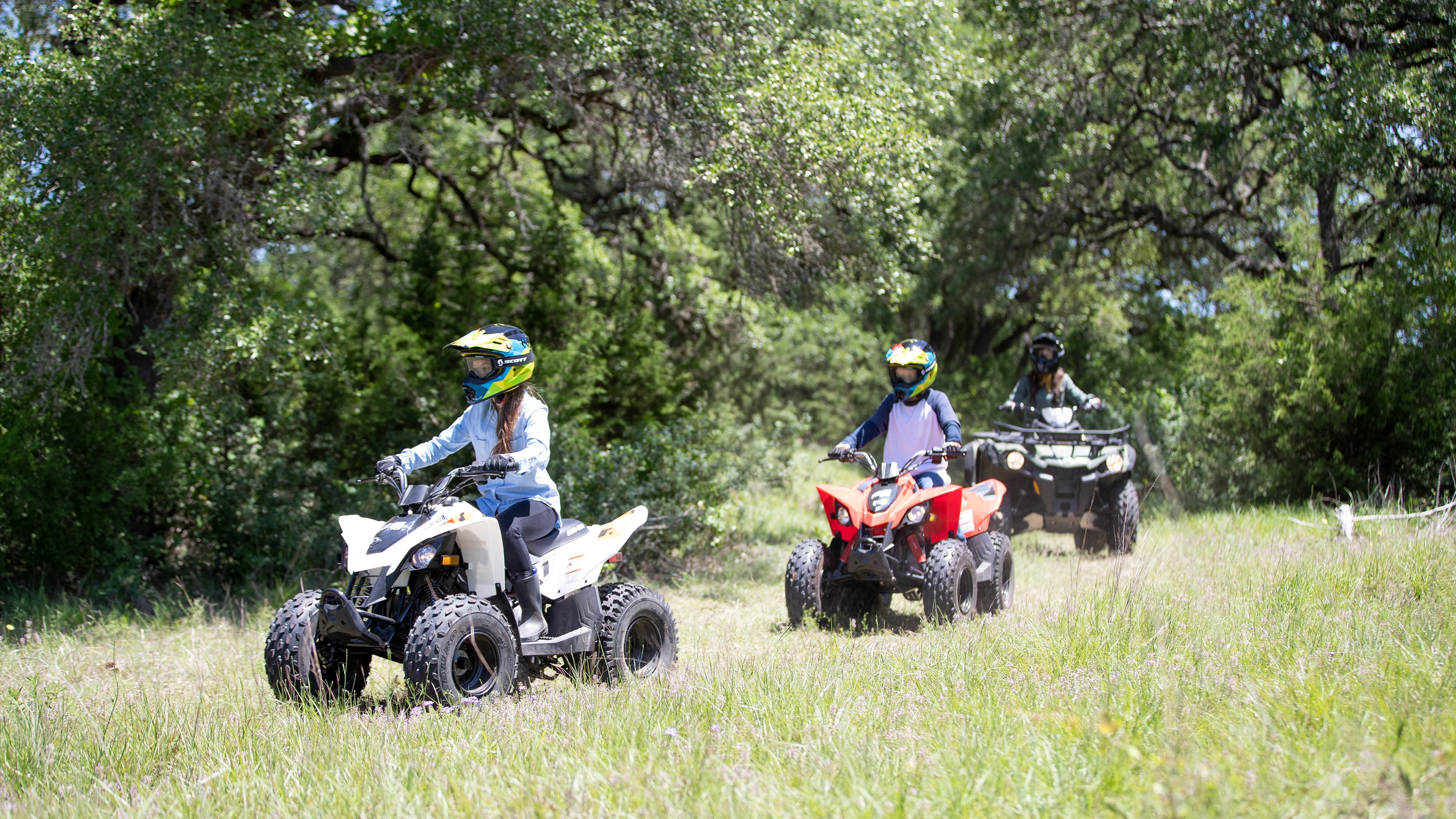 Two kids riding Can-Am DS ATVs and an adult in the background