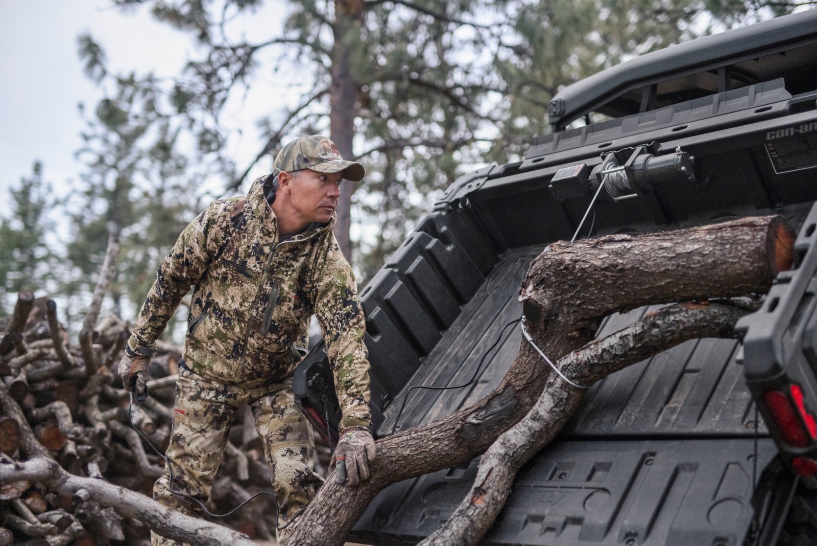 Un homme habillé en camouflage utilisant le treuil dans la boîte de son Can-Am Defender pour charger du bois
