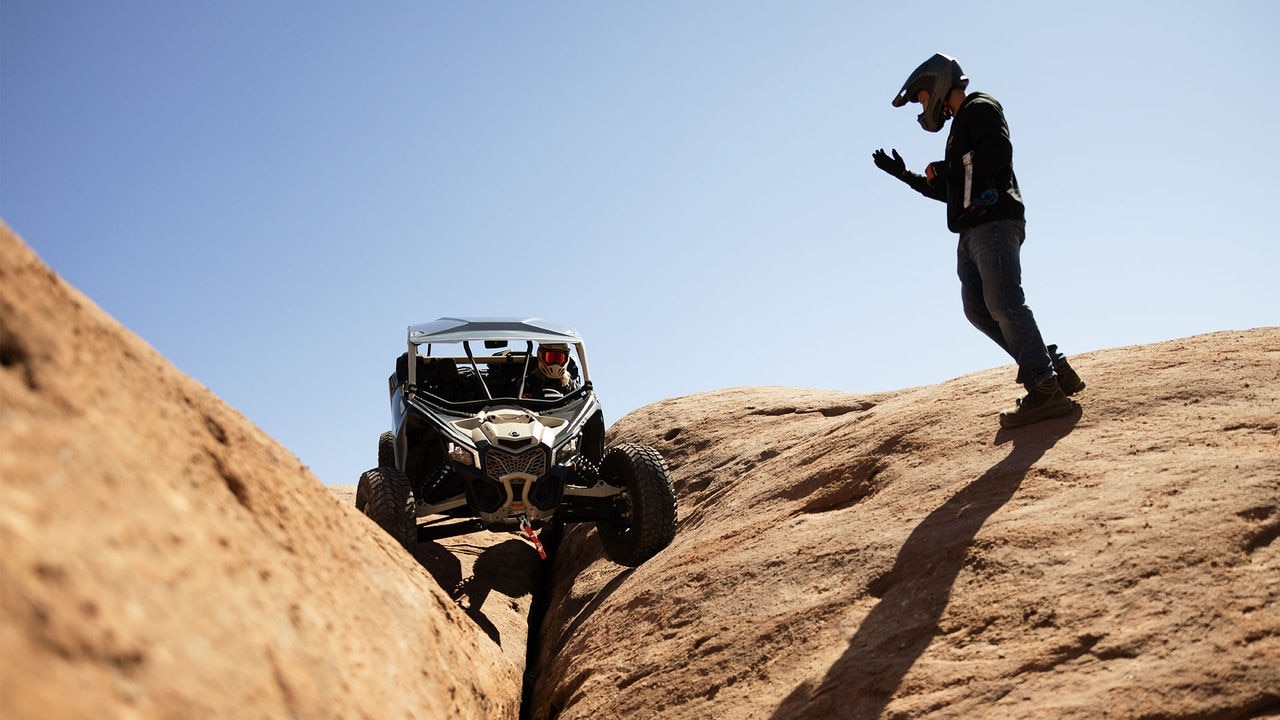 Conducteur avec casque Can-Am penché vers l’avant pour regarder un autre conducteur de Can-Am Maverick sur des roches.