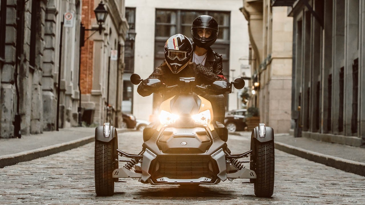 Una pareja conduciendo su Can-Am Ryker con paneles en amarillo deslumbrante por una calle adoquinada