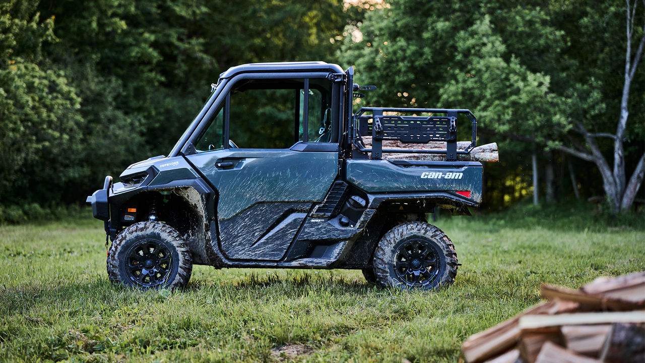 Un véhicule côte à côte Can-Am Defender XU HD10-HD11 2027 sur un vaste terrain, avec du bois dans sa caisse de chargement
