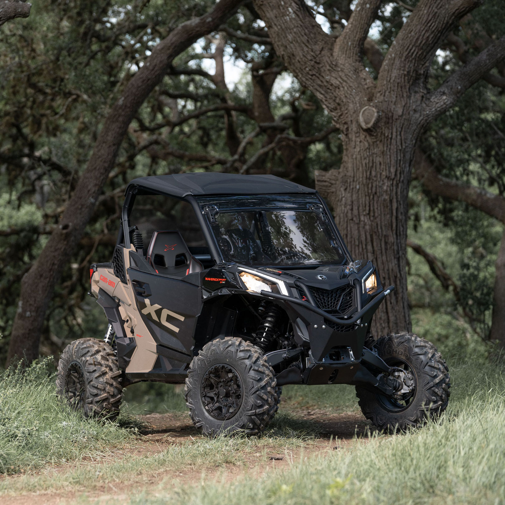 Een Desert Tan en Carbon Black Can-Am Maverick Sport X XC geparkeerd in een bosrijke omgeving 