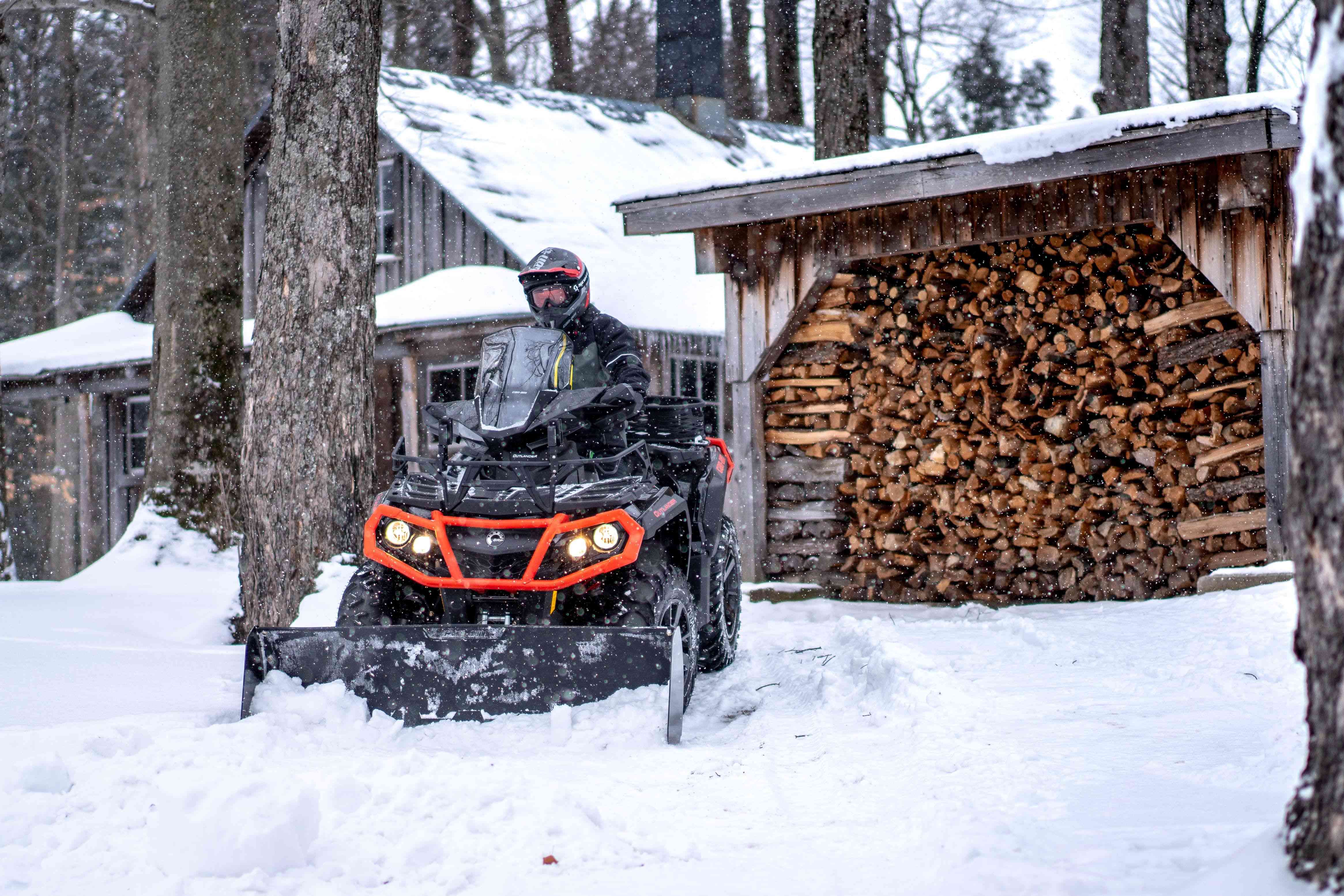 Man plowing snow with a Can-Am Outlander ATV near a wooden chalet