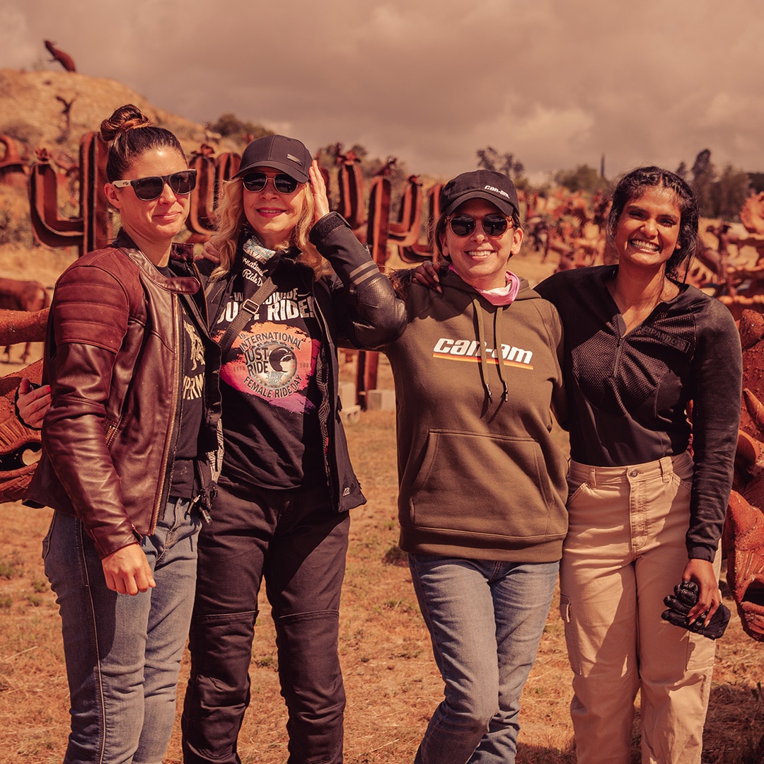 Four smiling women in motorcycle gear, in the desert in front of some cacti