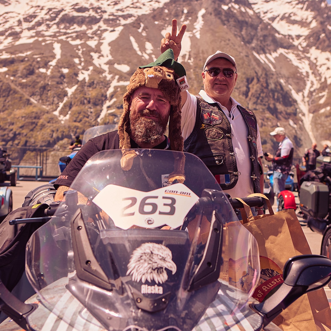 Two smiling men standing in front of a Can-Am three-wheeler against the backdrop of snow-capped mountains