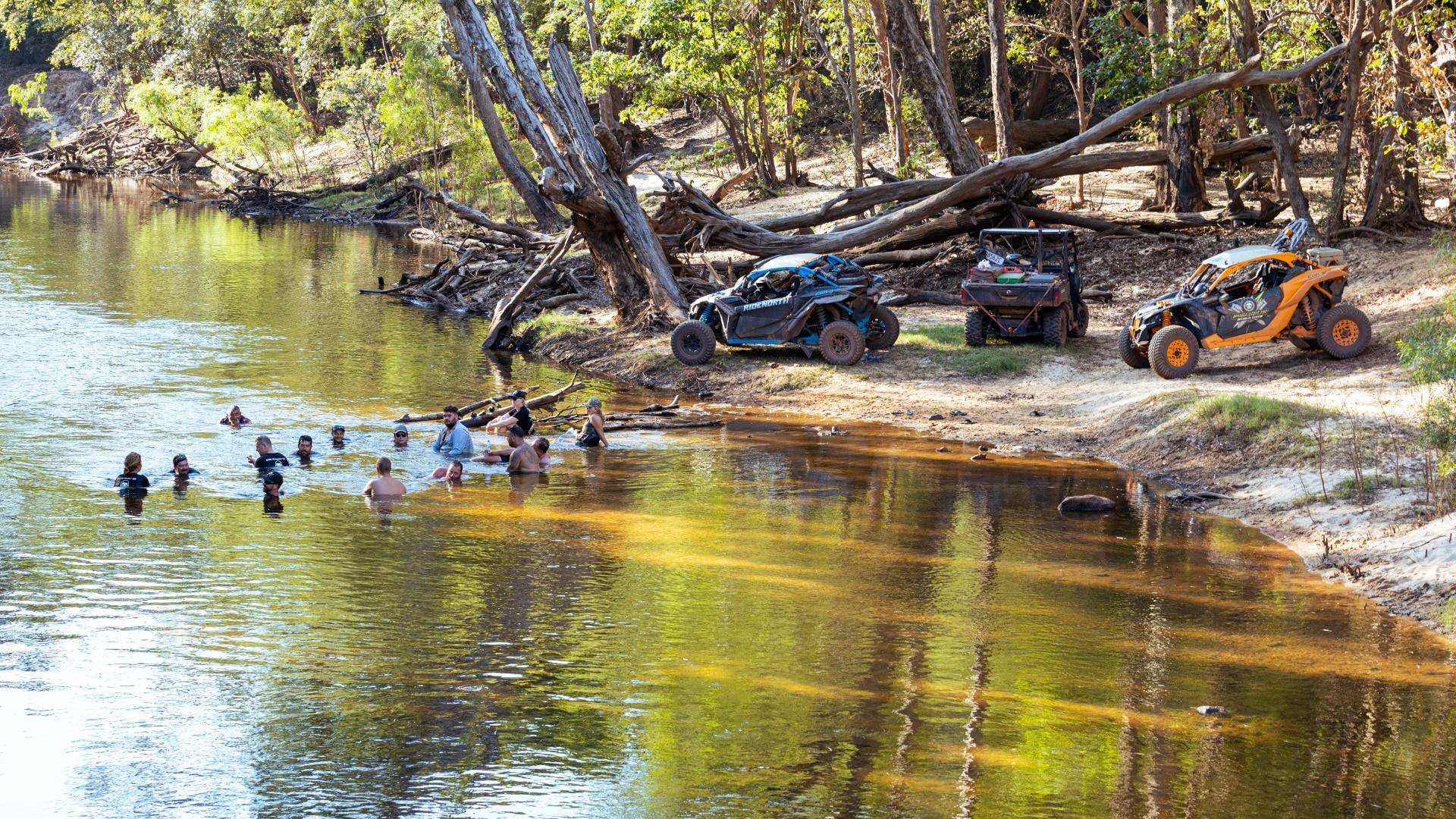 People swimming in a creek while their Can-Am units are parked on the sand bank. 