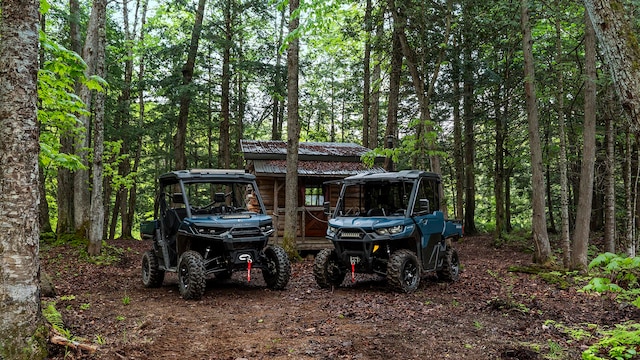 Two 2026 Can-Am Defender parked in a forest near a cabin