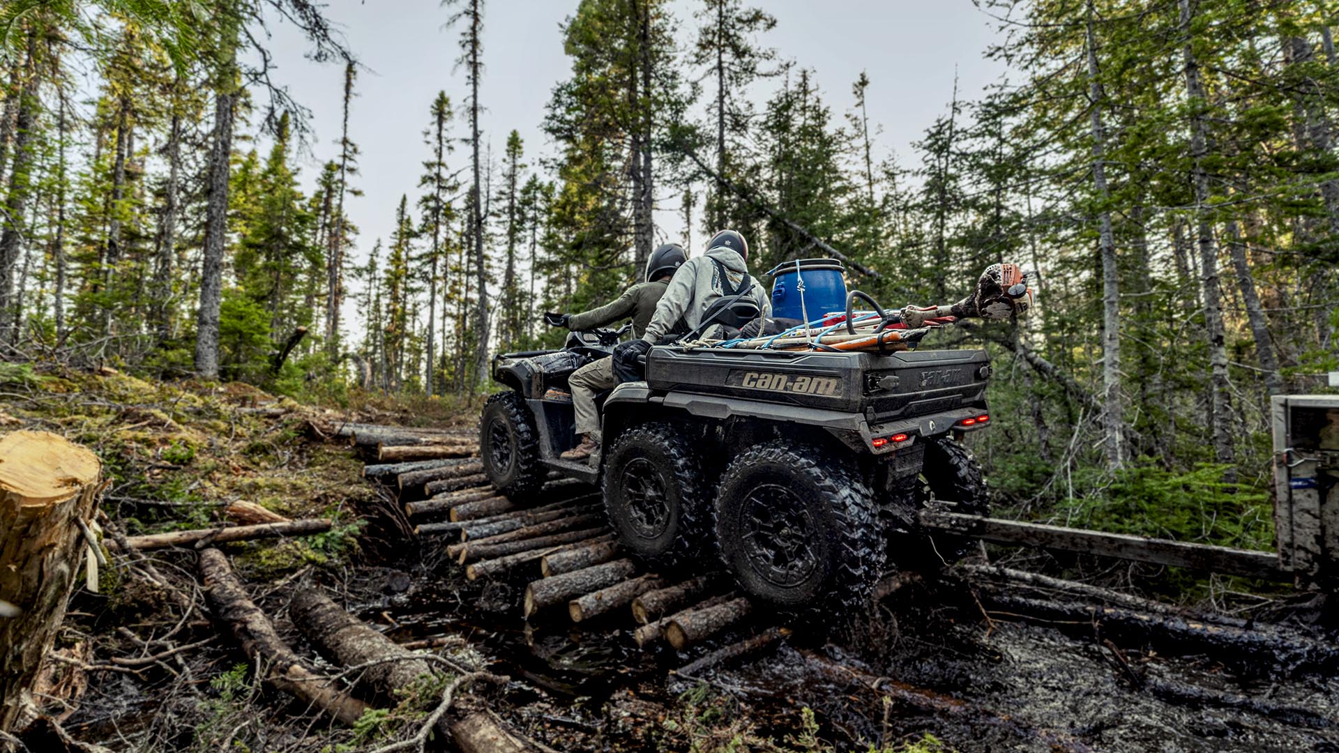 Two people on a Can-Am Outlander MAX 6x6 ATV riding over tree trunks in the middle of the forest