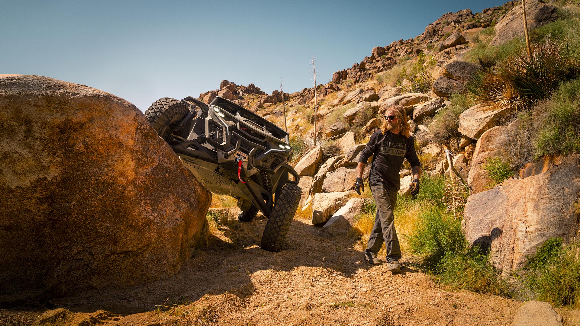 A Can-Am Maverick X RC climbing a rock, with a rider walking alongside, in the desert