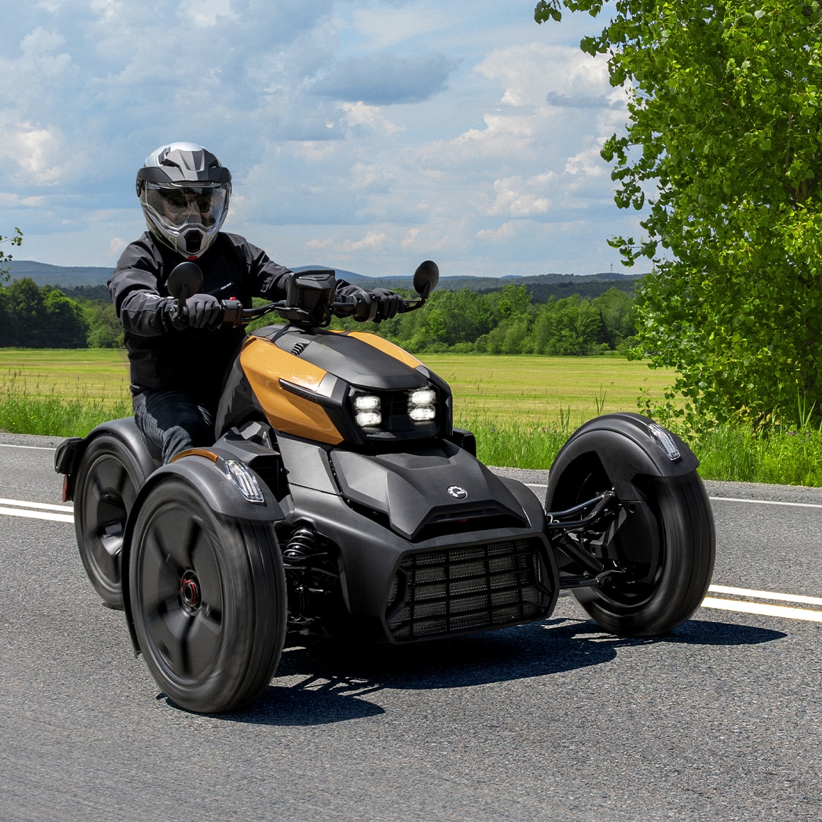 Motocycliste conduisant une moto 3-roues Can-Am Ryker 2027 sur une route pavée à côté d'un champ