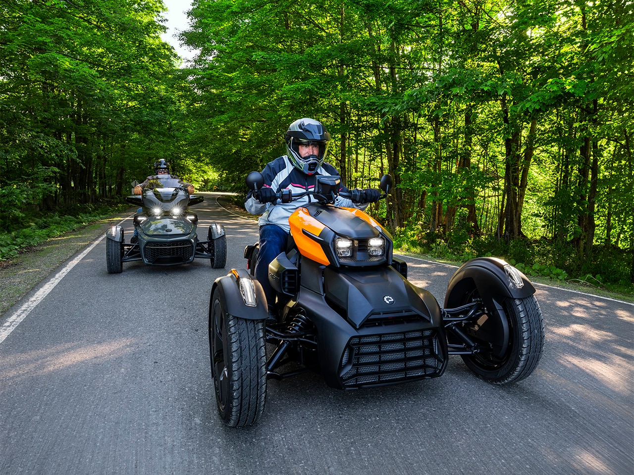 Un Can-Am Ryker 2027 suivi d’un Can-Am Spyder 2027 sur une jolie route bordée d’arbres