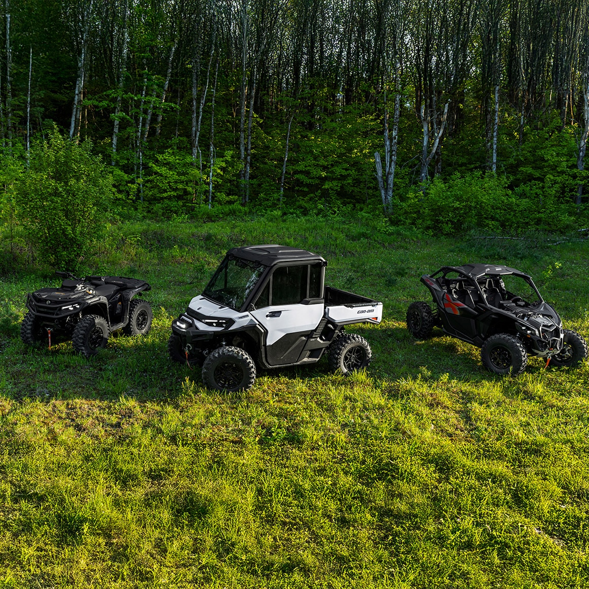 Two Can-Am side-by-side vehicles and an ATV parked in the grass near a forest