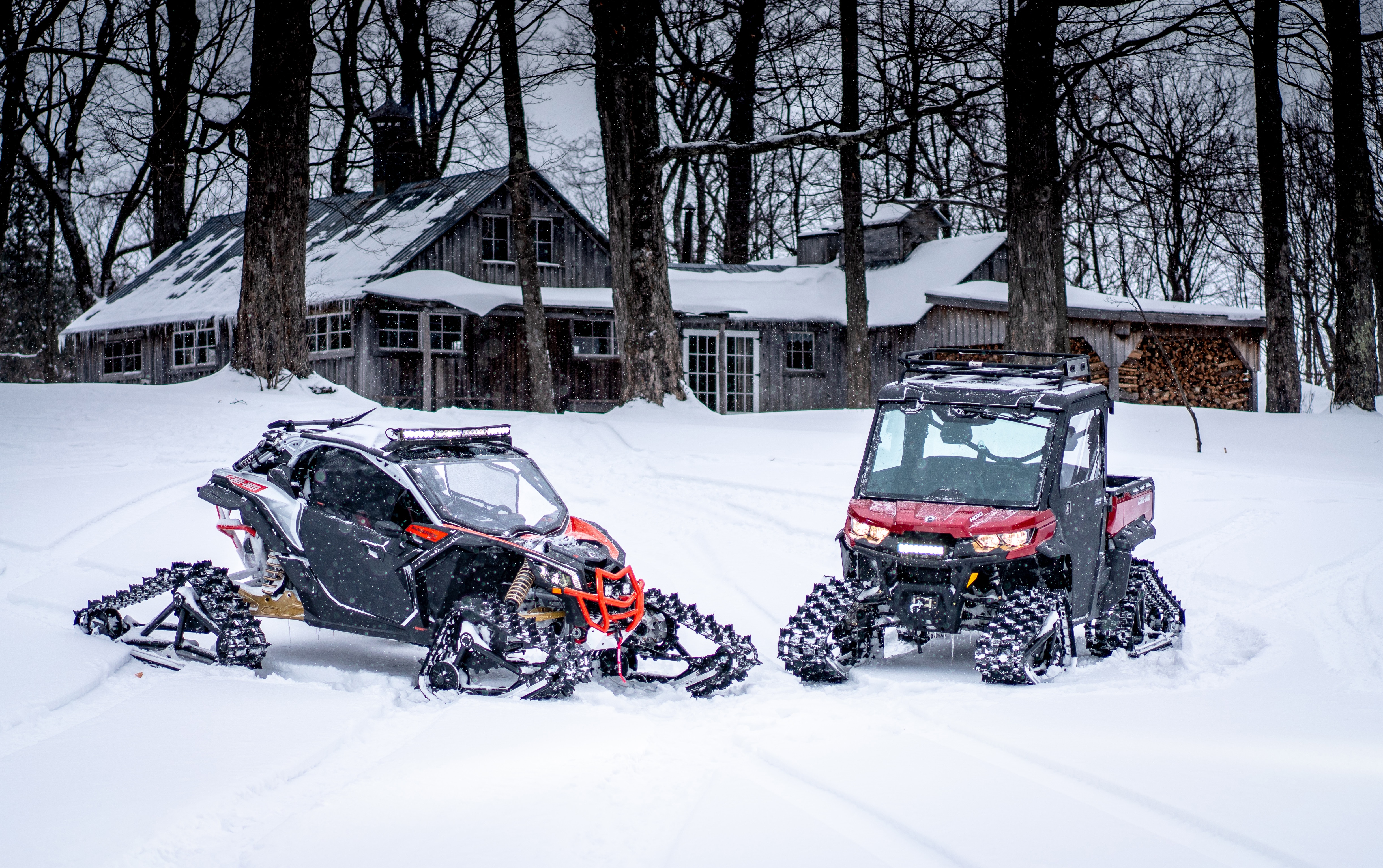 A Can-Am Maverick X3 and a Defender with track kits in the snow near a wooden chalet