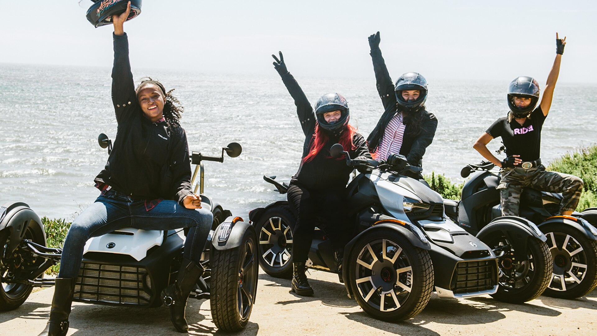 Group of women sitting on their Can-Am Ryker with raised arms in front of the sea.