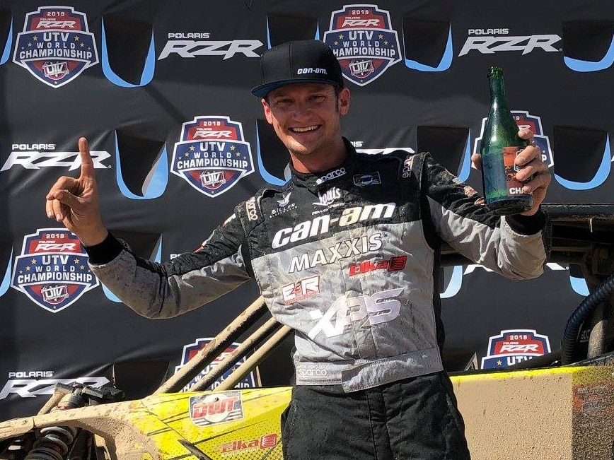 Cody Miller on a podium, holding a champagne bottle and his index finger as a "number one" sign. 