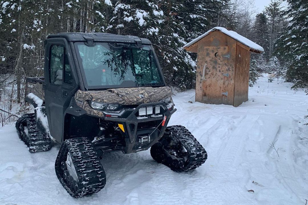 A Can-Am Defender Limited with track systems on snow near a small outdoor wooden toilet