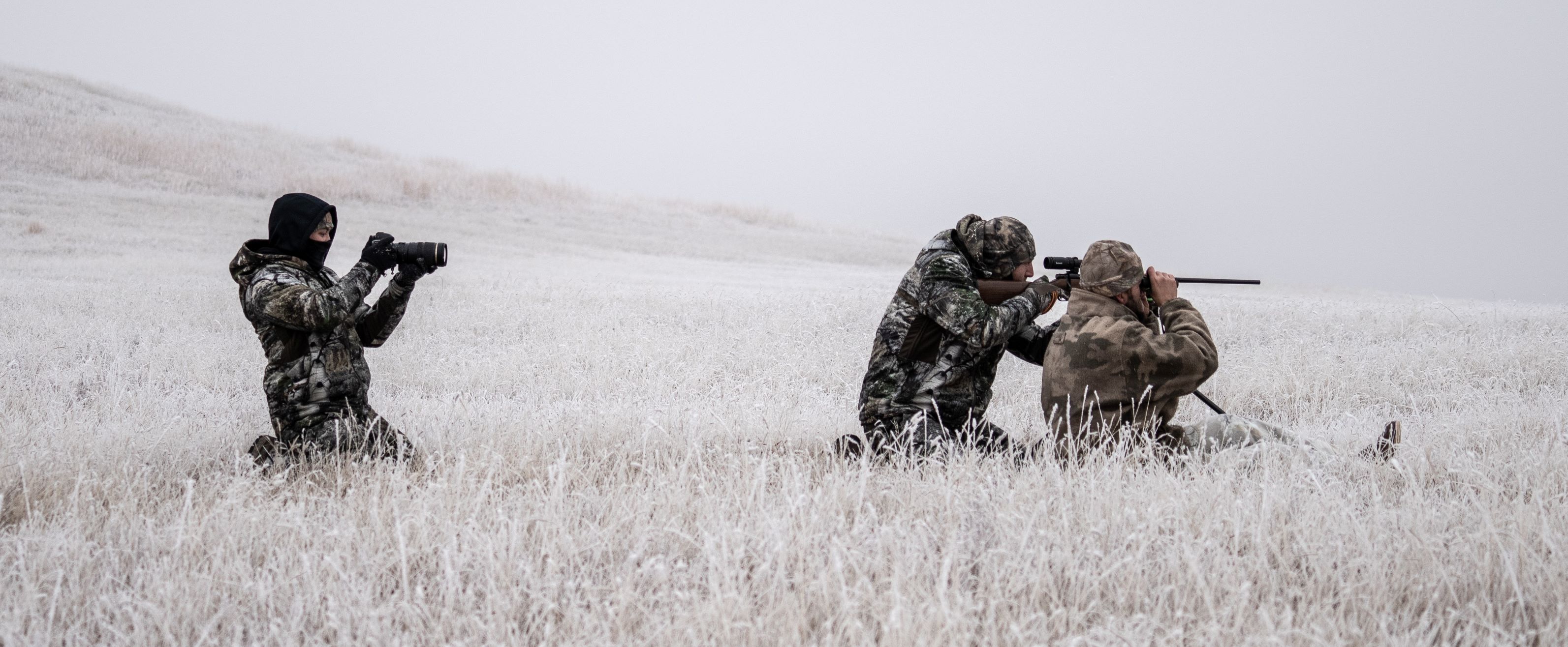 A hunter kneeling in a foggy field in shooting position with two other hunters.