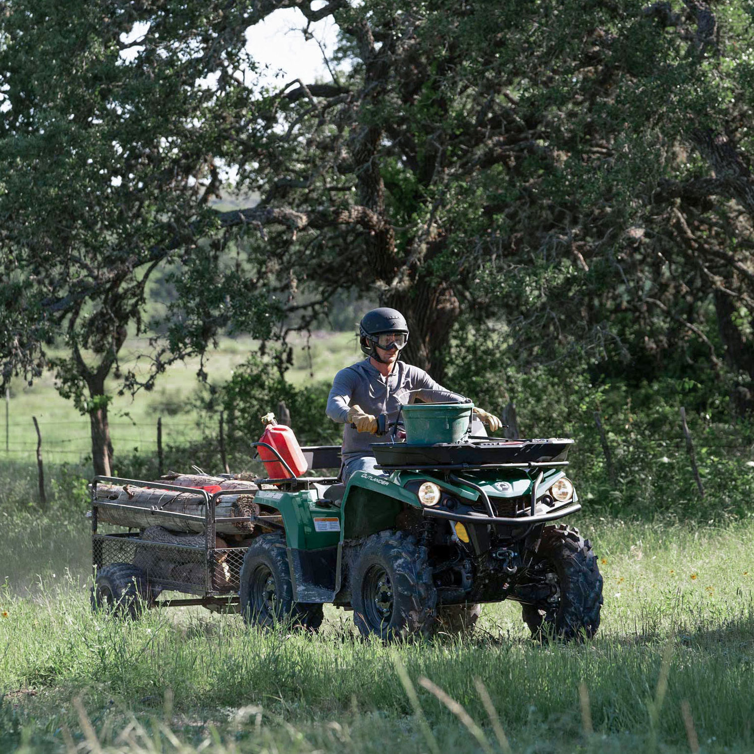 Man riding a tundra green Can-Am Outlander ATV with a trailer filled with woods