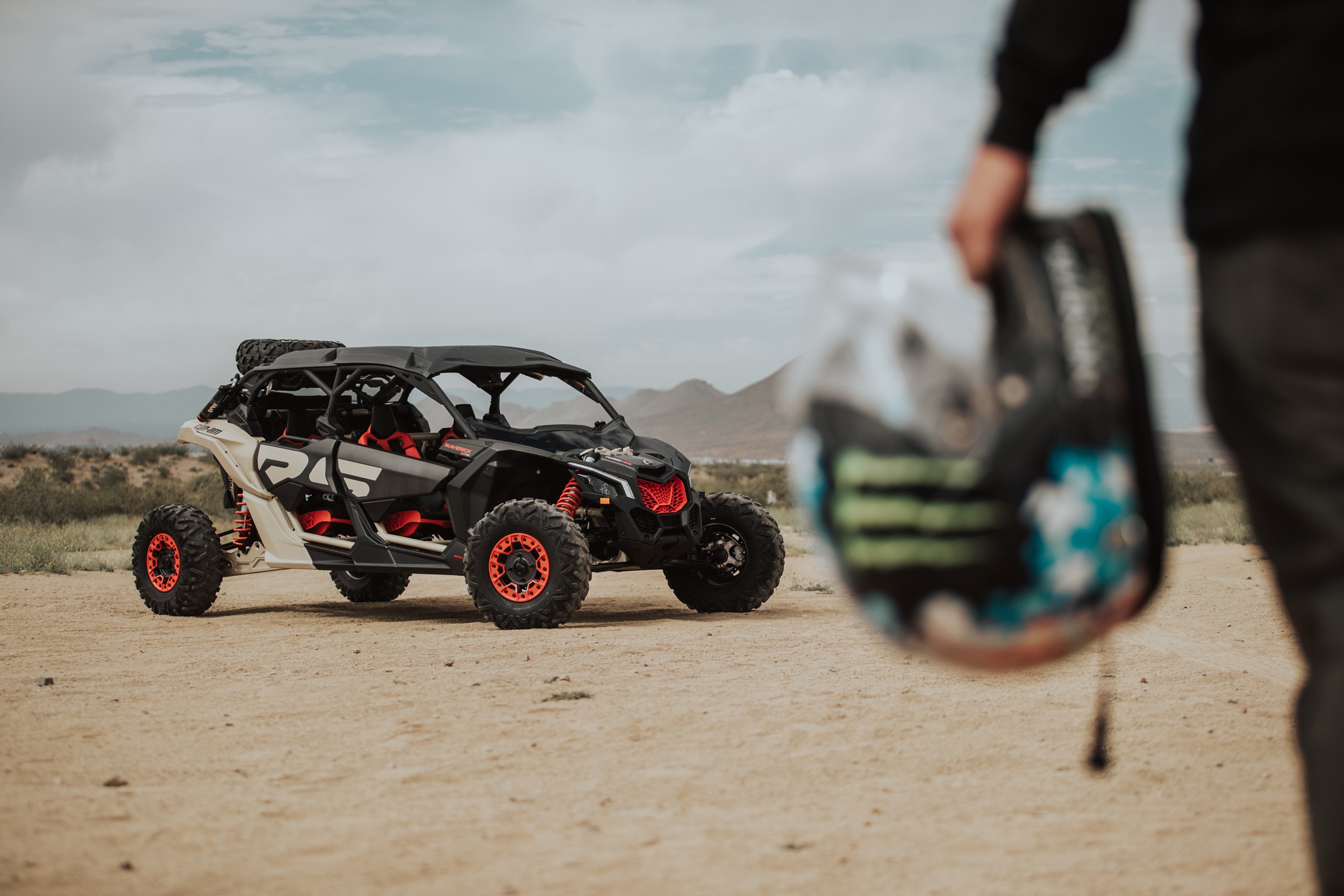 Can-Am Maverick MAX X3 X rs Turbo RR parked on the sand in front of mountains with the rider holding his helmet in the foreground