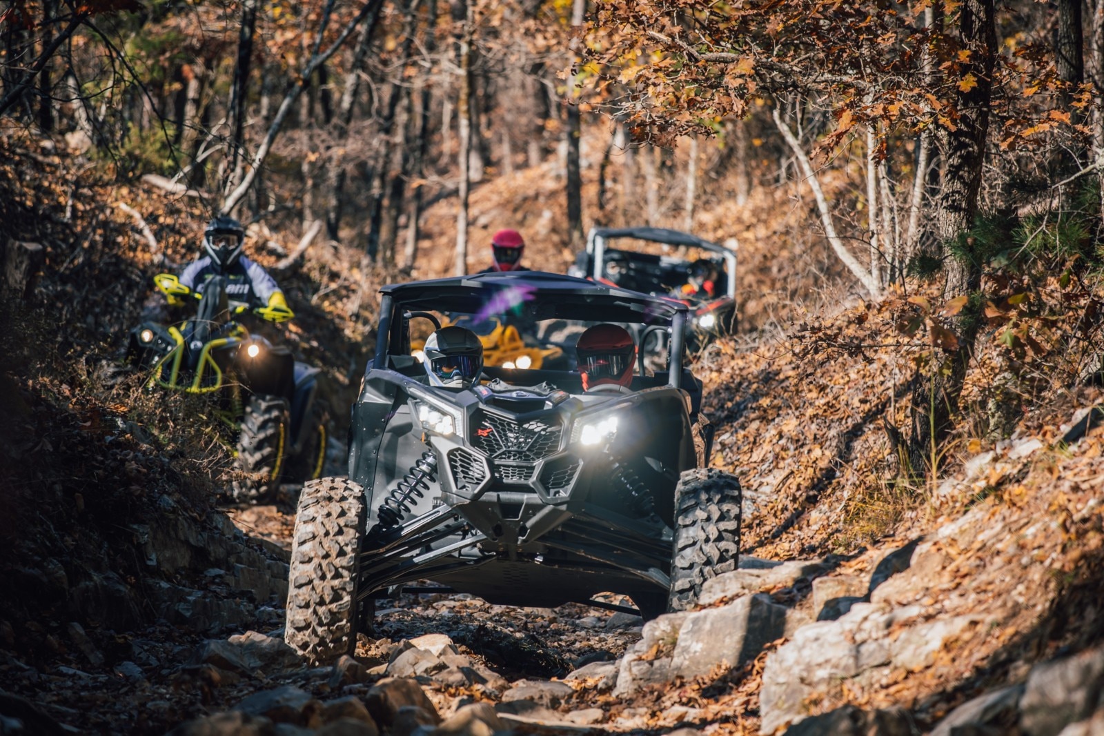 Two riders in a Can-Am side-by-side, on a rocky trail in the woods with other riders.