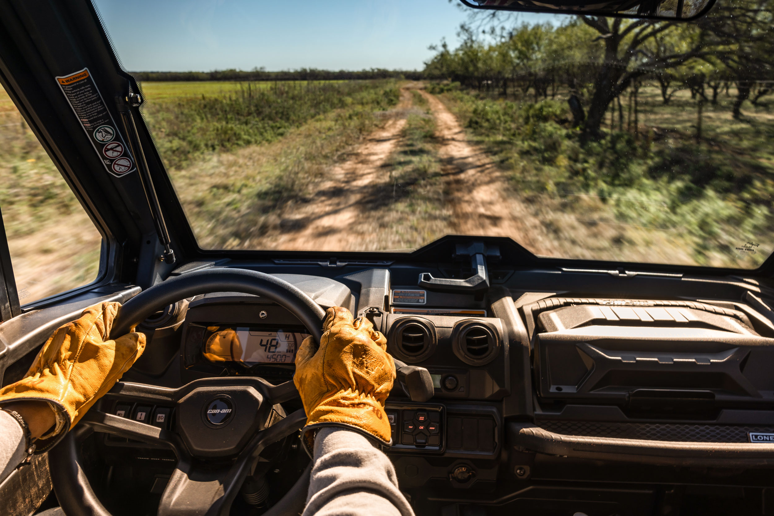 Can-Am driver's hands on the wheel, from inside the cab, driving down a dirt road on a farm.