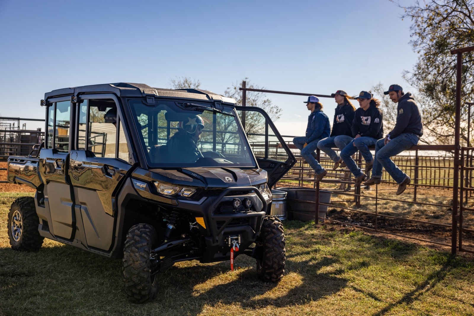 Four ranchers sitting on a cattle fence next to a parked Night Black Can-Am Defender MAX Lonestar CAB