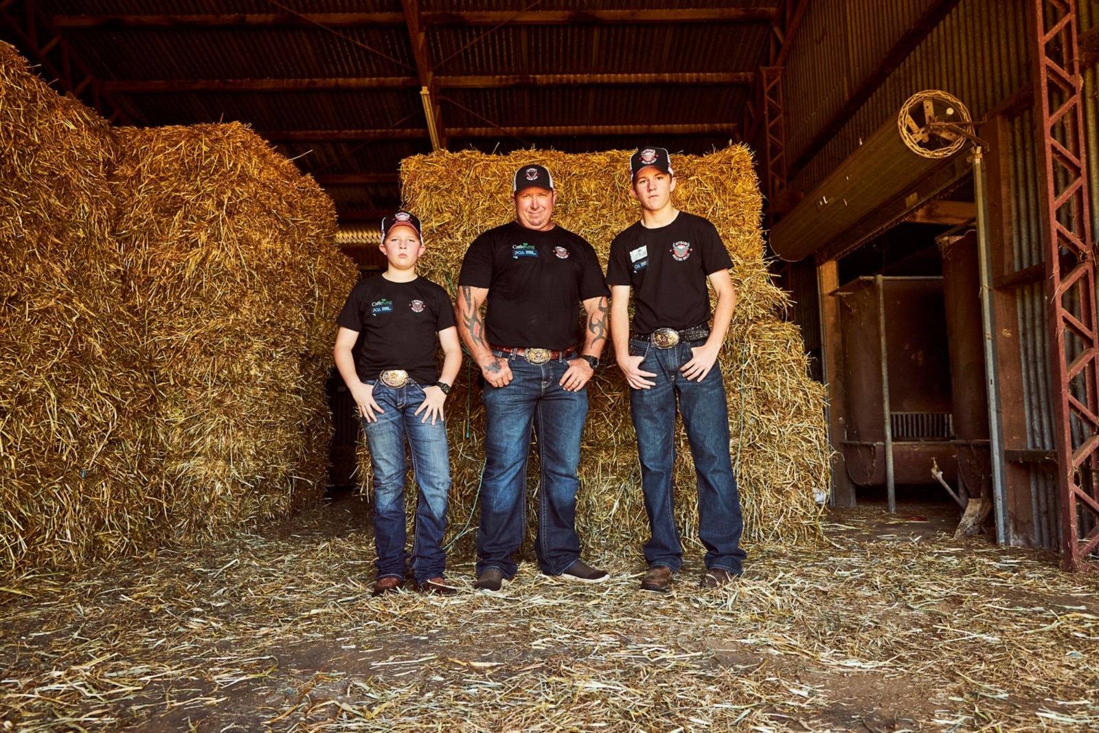 Jason Dittman and his two eldest sons, repping their belt buckles and denim, standing in front of square hay bales