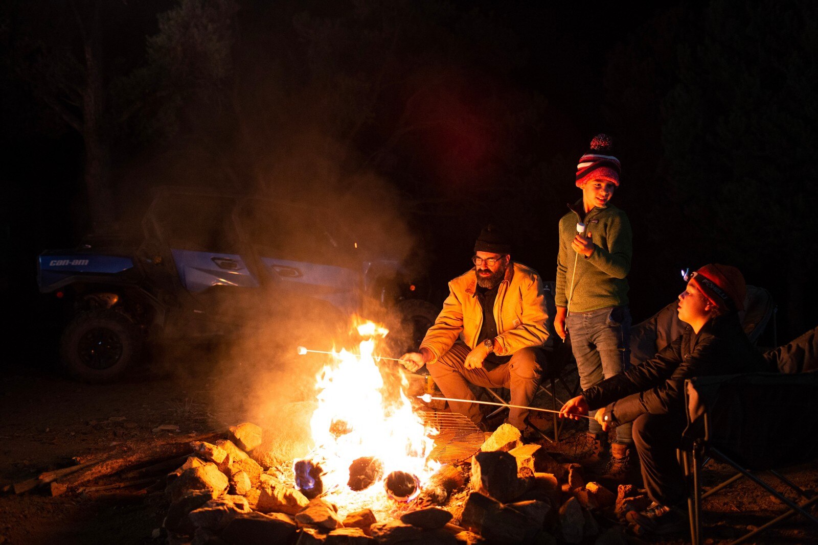 Foto nocturna de Billy Parks y sus dos hijos asando malvaviscos en una fogata con su Oxford Blue Can-Am Commander MAX XT estacionado en el fondo