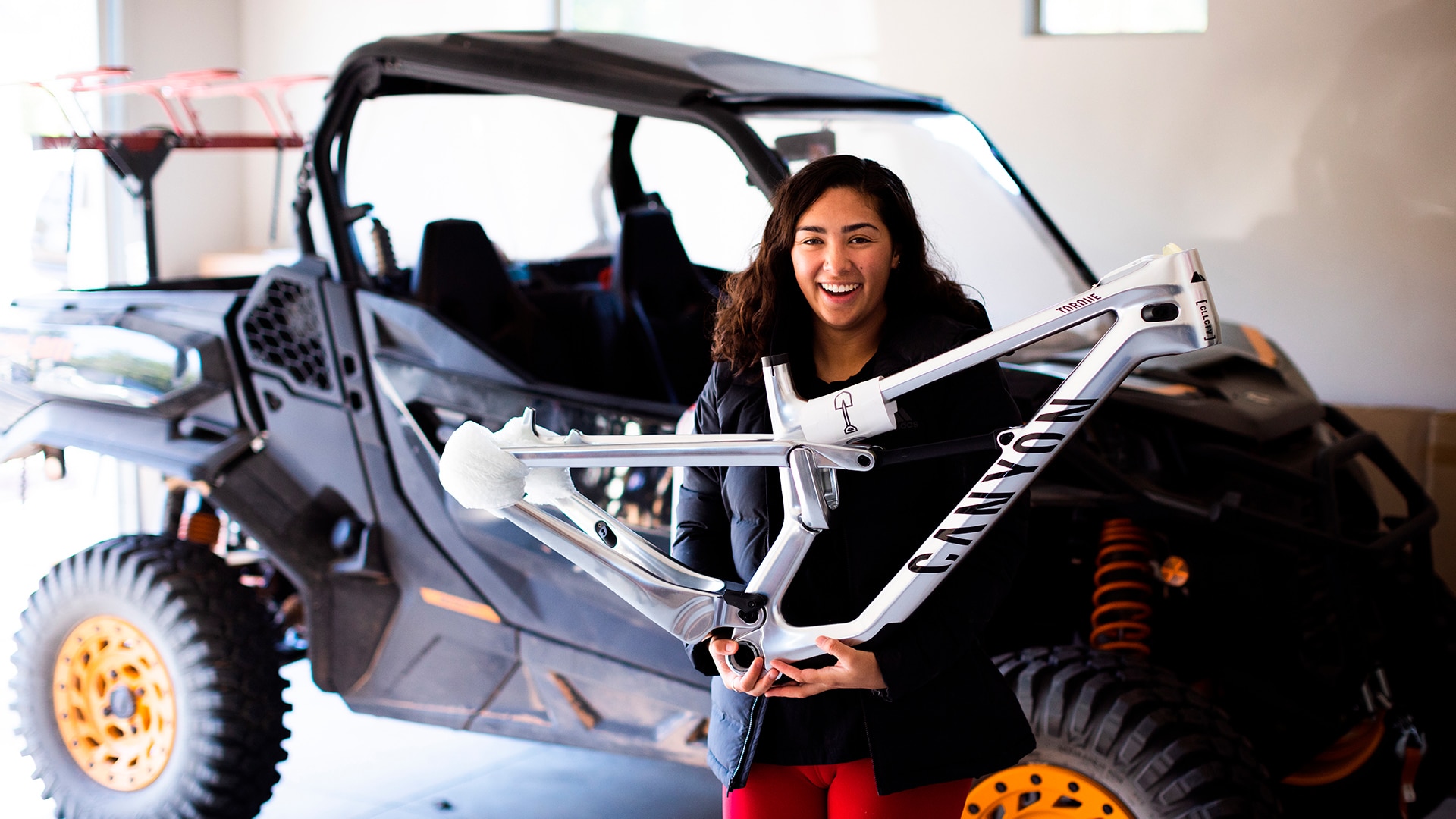 Samantha Soriano sonriendo, apoyada en un Can-Am Commander con detalles en naranja y sosteniendo un cuadro de bicicleta