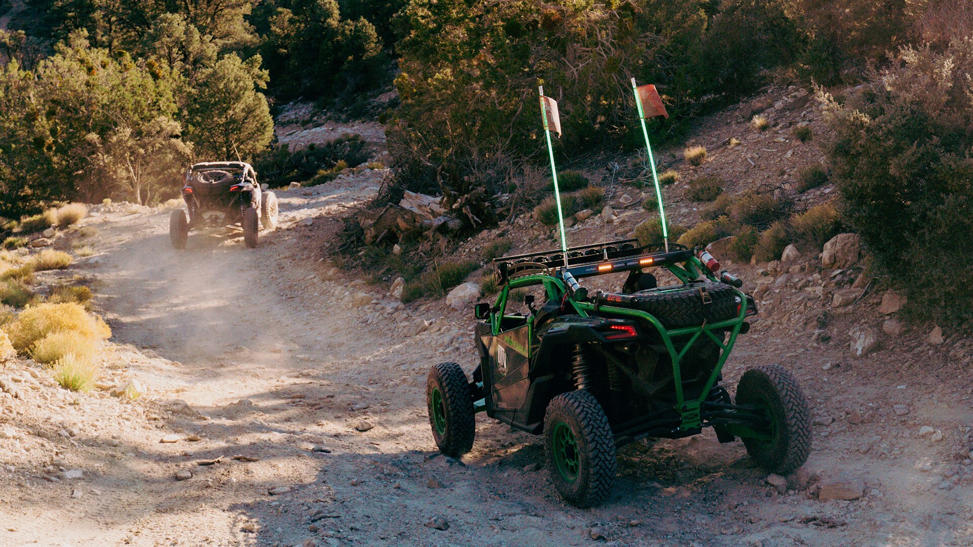 Can-Am riders driving side-by-side vehicles on a dirt of trail leading to a forest.