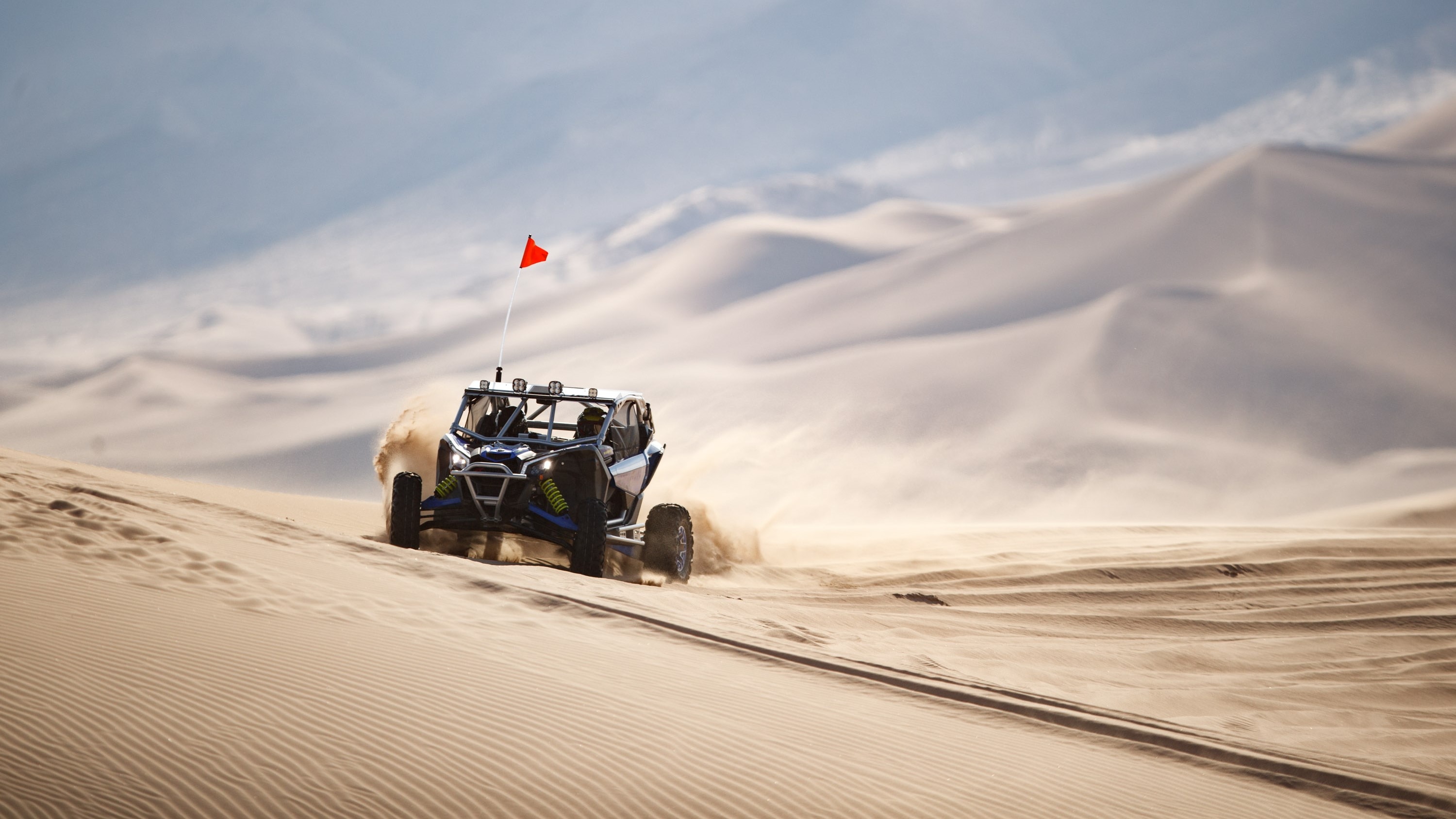 Side view of a Can-Am Maverick X3 kicking up sand as it drives at the base of a rock pile. 