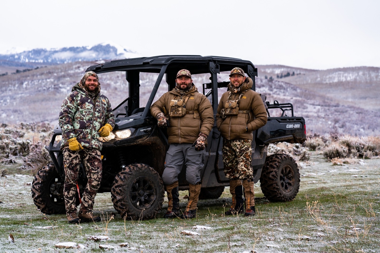 Three hunters from the Hushin' crew dressed in camo, leaning on their Defender parked in a field at the base of snowy mountains.  