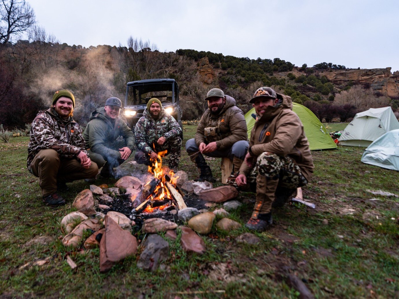 5 members of the Hushin' crew around a camp fire with their Defender and tents set up in the background. 