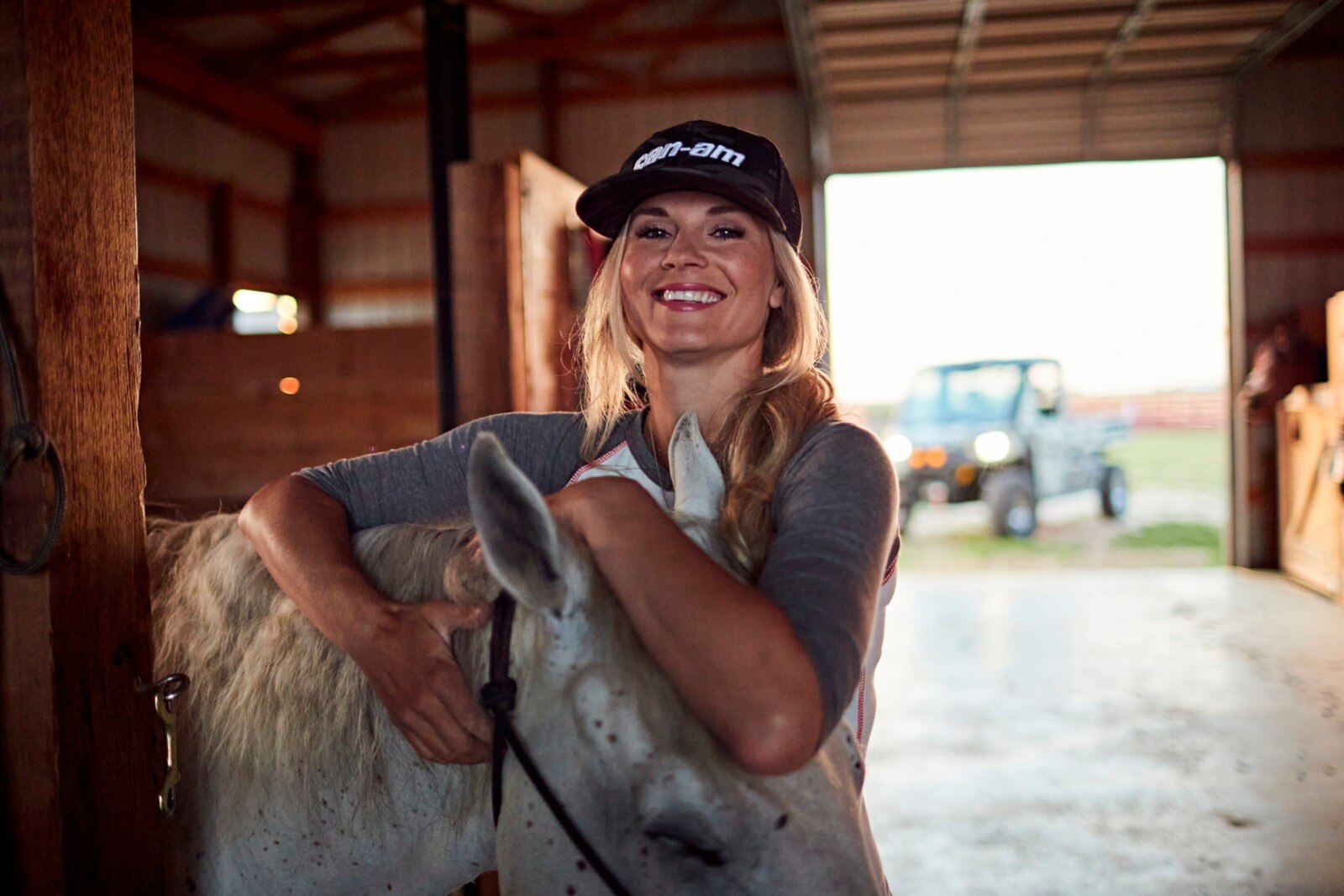 Kristy Lee Cook wearing a Can-Am cap, holding her horse's head in her arms, in her barn with her Defender in the background. 