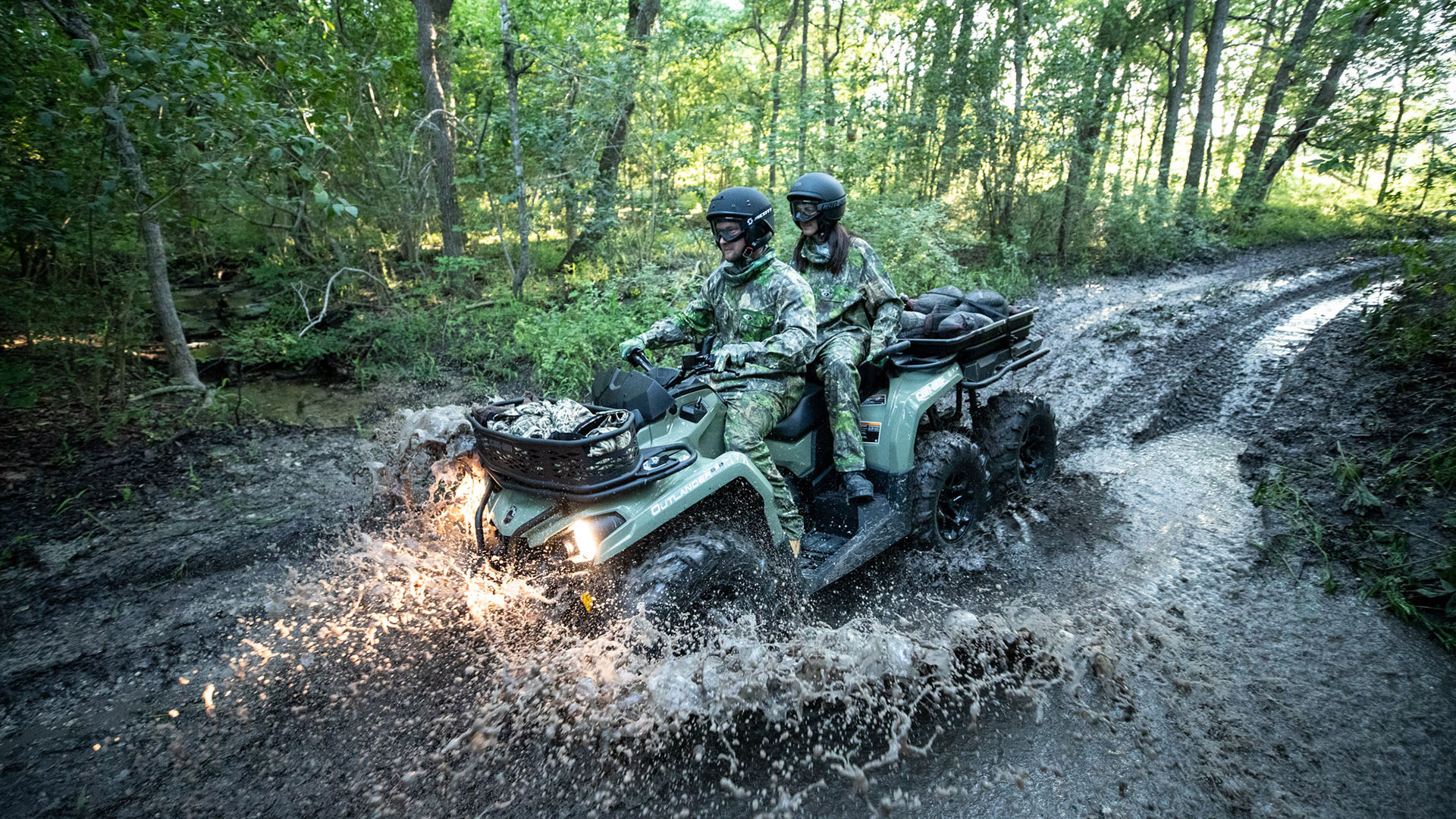Two riders in camo on a Can-Am Outlander 6x6 MAX driving through a mud hole 