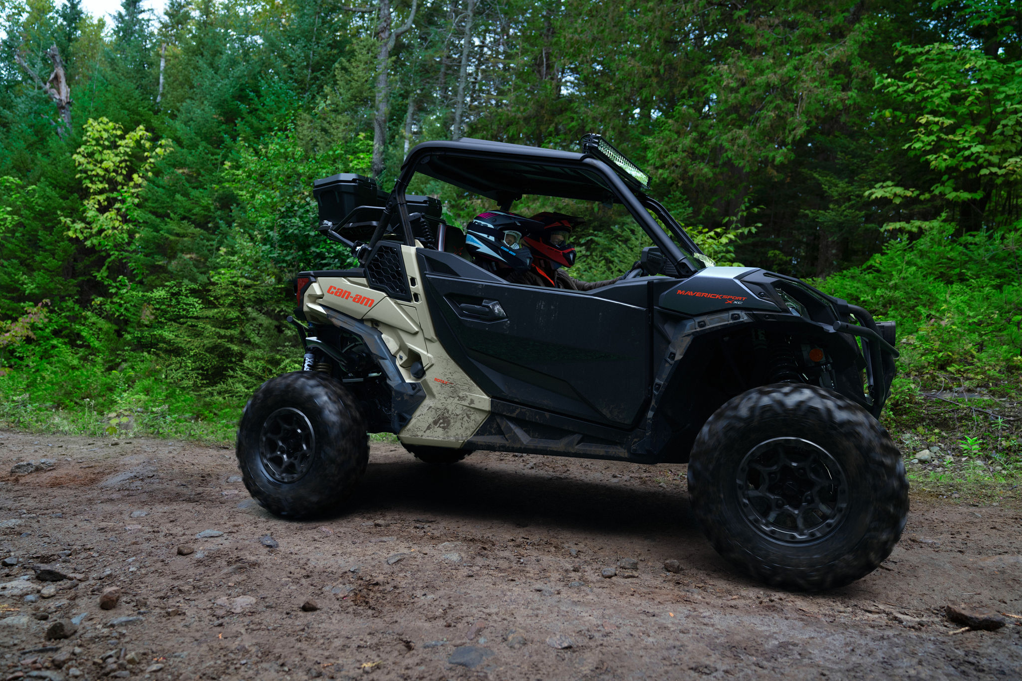 Two riders in a desert tan and carbon black Can-Am Maverick Sport X XC on a trail