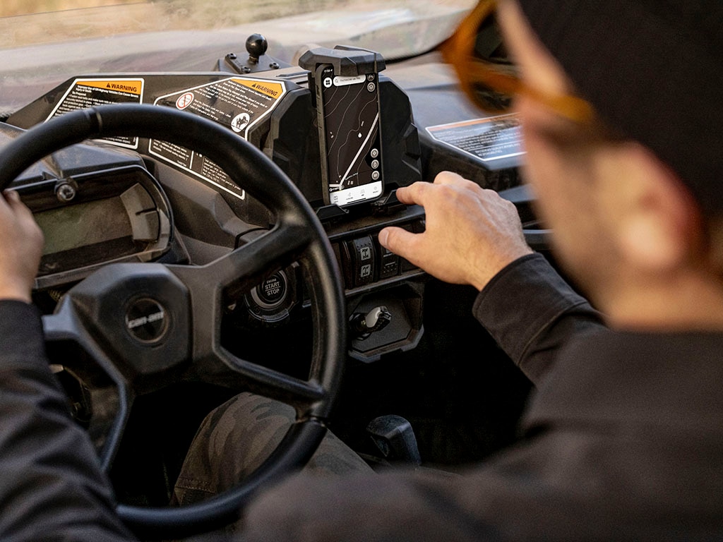 Primer plano desde atrás de un conductor al volante de un Can-Am SSV, con la mano derecha cerca del dispositivo en el soporte del smartphone.