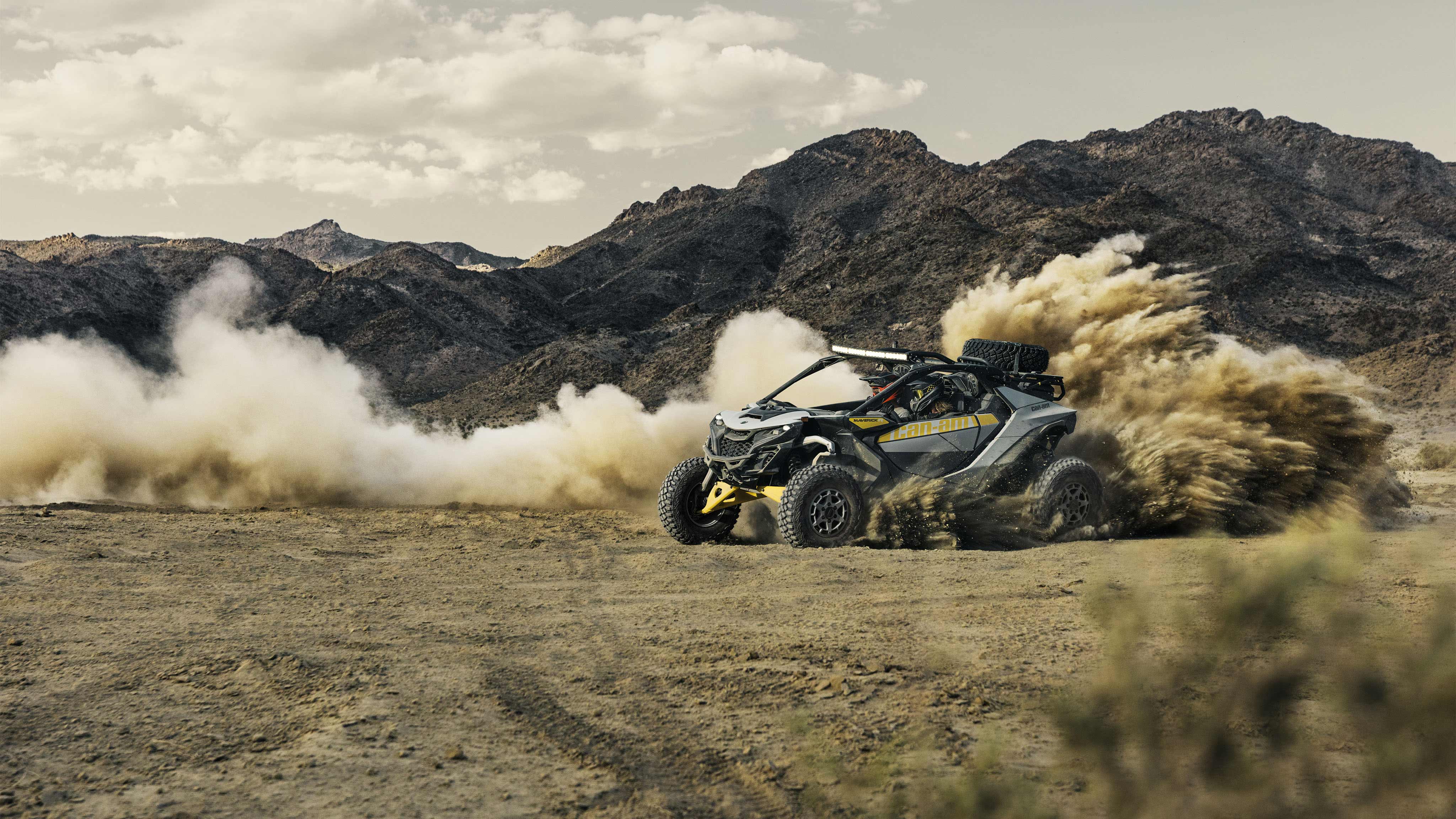Two riders in a Can-Am Maverick R making a hard turn on a dirt road