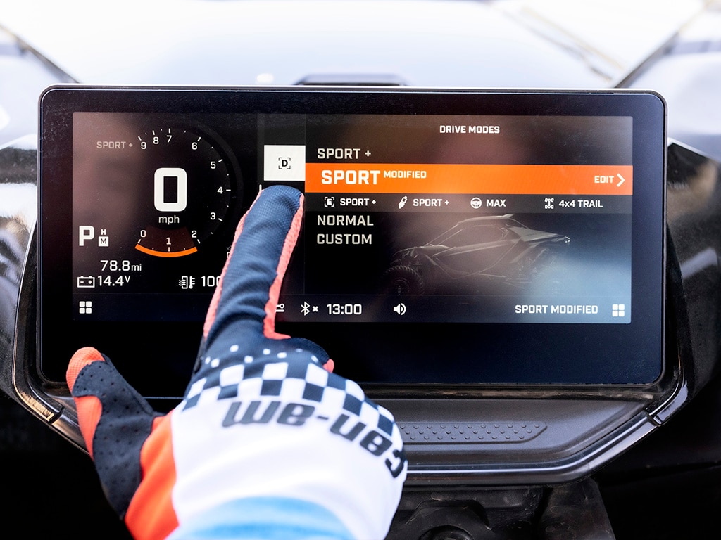 A hand wearing a colourful Can-Am glove is tapping a drive mode option on the touchscreen using the right index finger