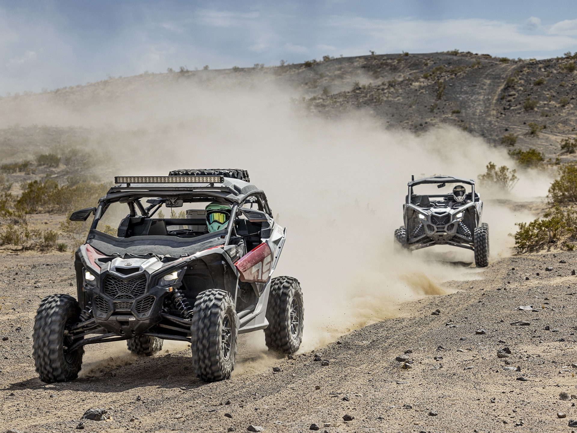 Two Can-Am side-by-side vehicles riding on a dirt road