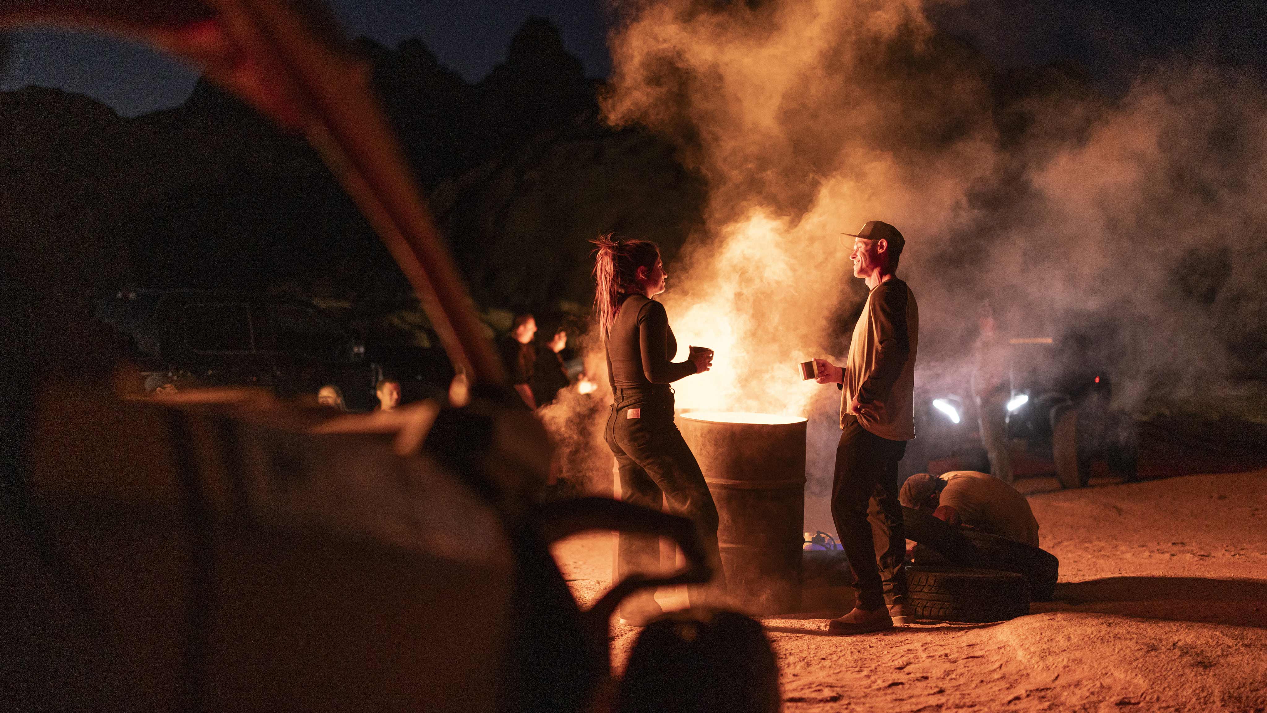 Dos personas charlan por la noche junto a un barril quemado en el desierto con un Can-Am Maverick R en primer plano