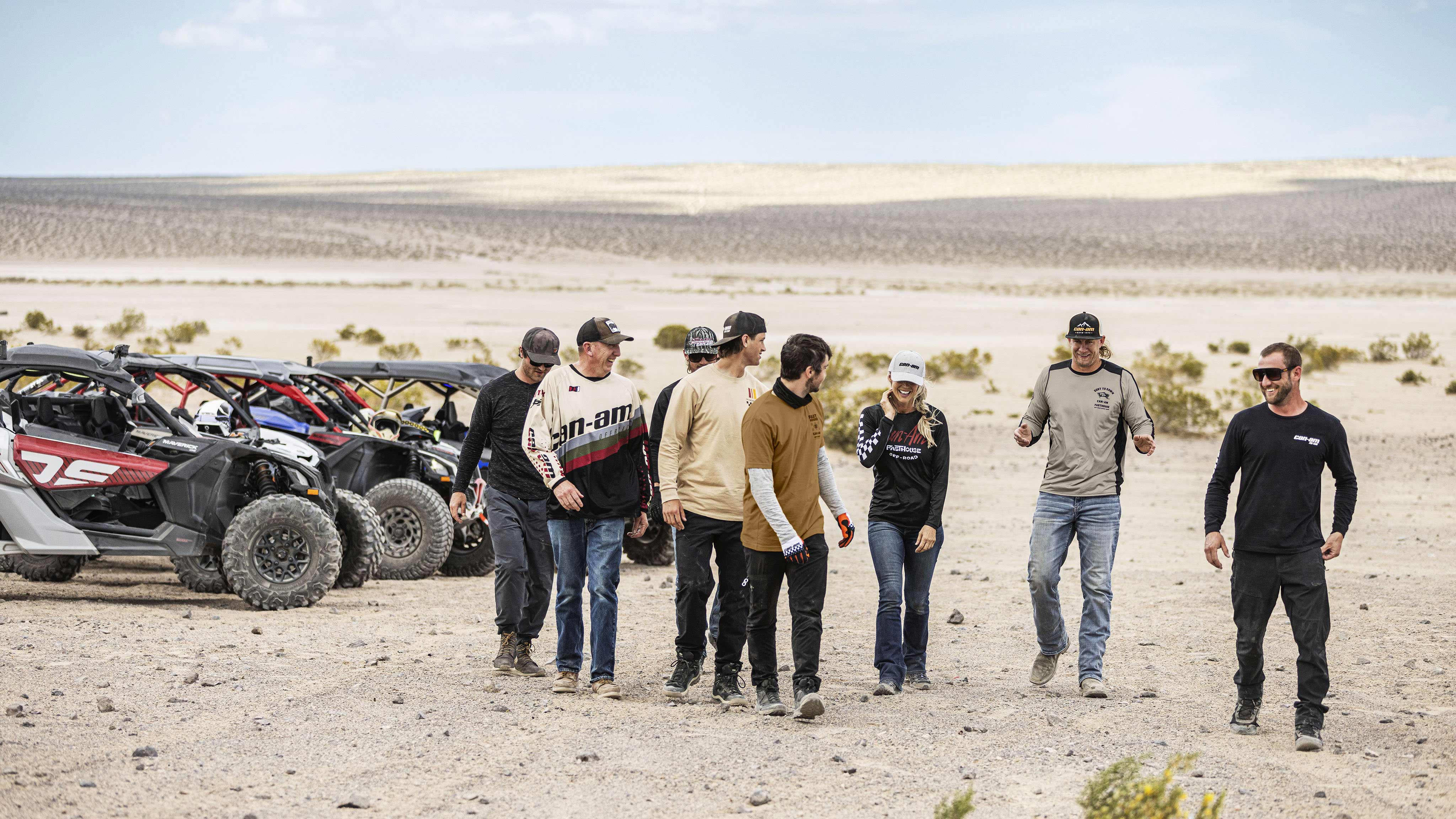 Oito pilotos caminhando em frente no deserto, com três veículos Can-Am Maverick ao fundo.